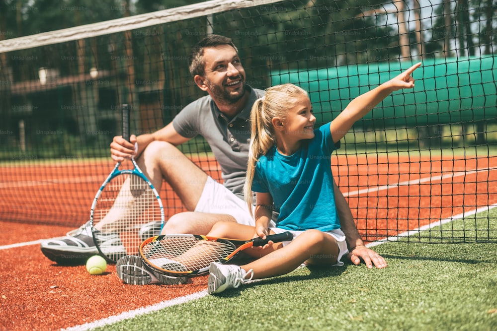 Cheerful father and daughter leaning at the tennis net and looking away with smiles while both sitting on tennis court
