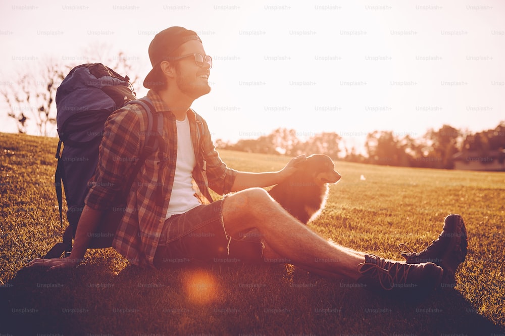 Cheerful young man with backpack petting dog while kneeling on the green grass outdoors