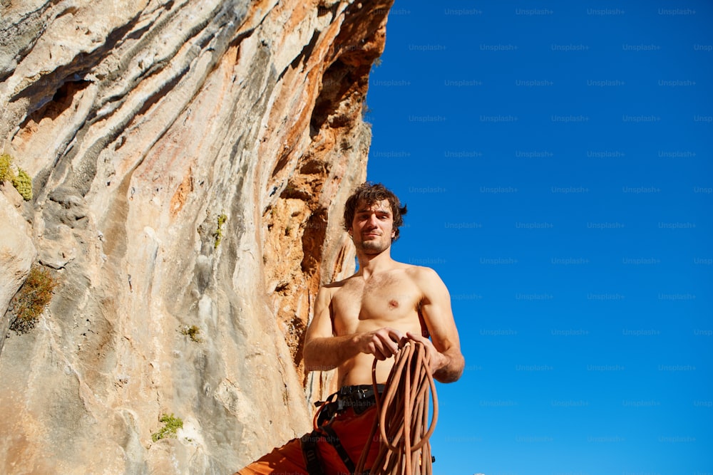  attractive muscular male climber with the rope under the cliff against blue sky
