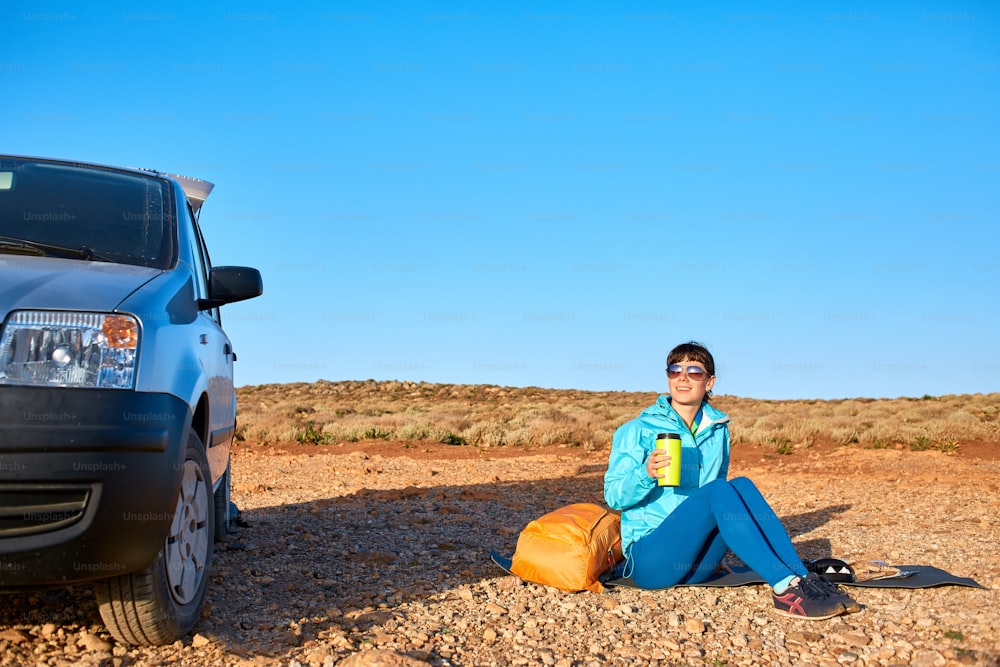 woman traveler sitting on the rug near the car and drink coffee. a woman in a place like a desert at dawn