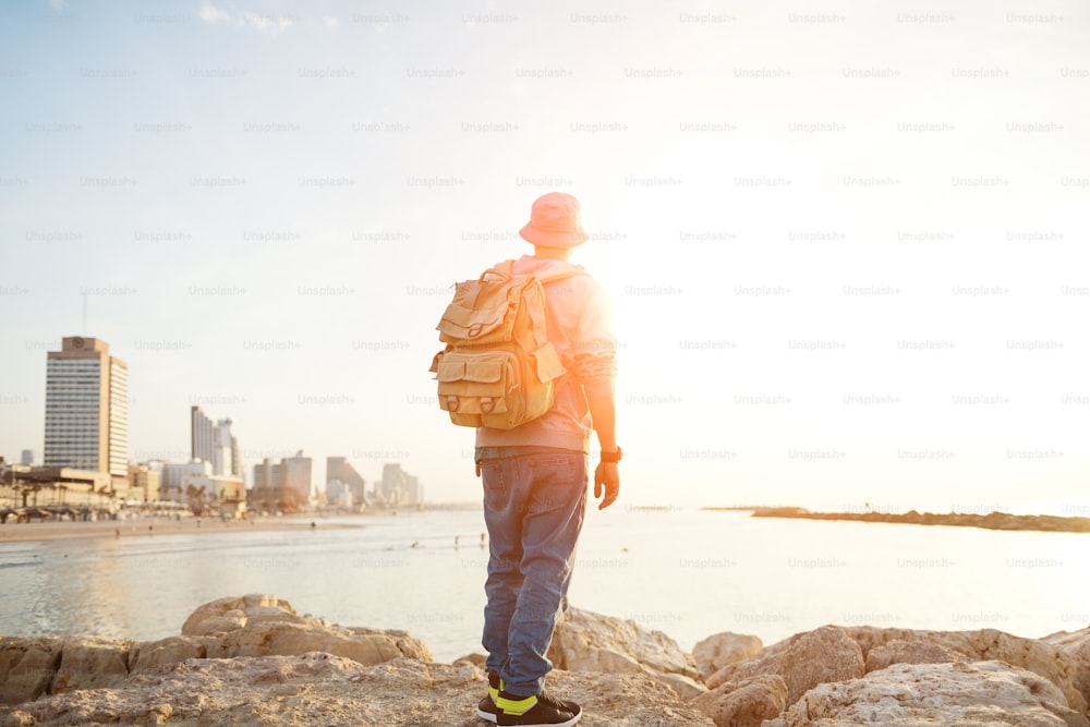 traveler with backpack standing on the rocks on the beach in the city and looking far away