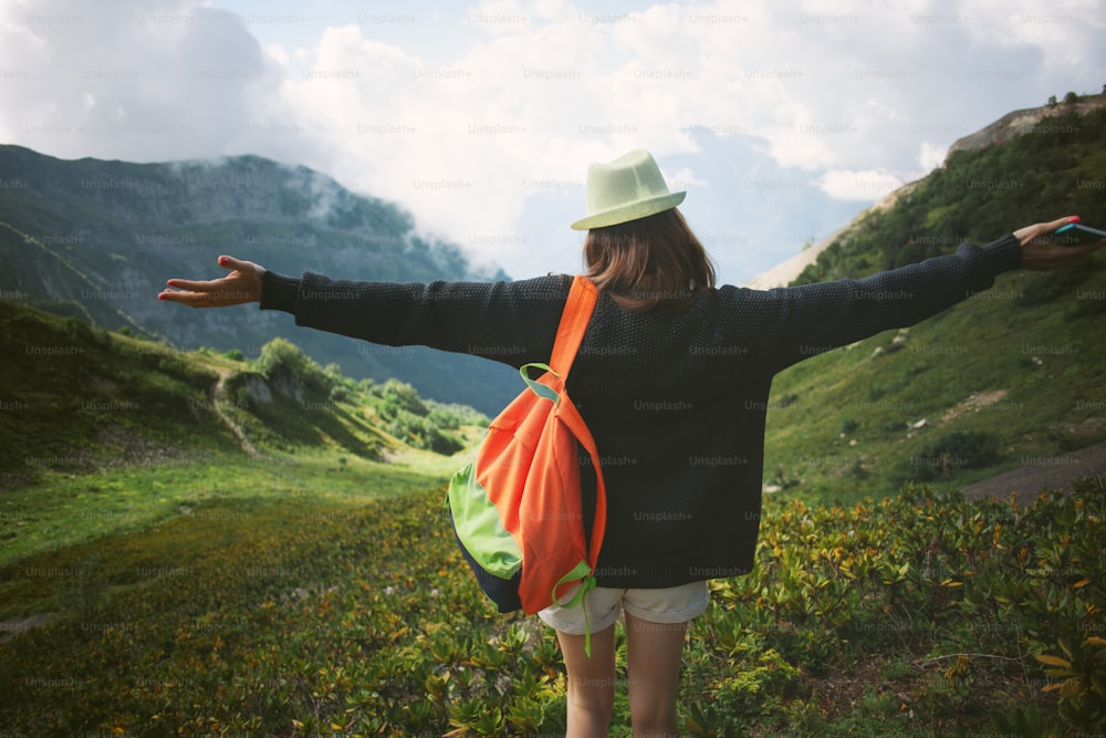 Young woman with outspread hands standing among beautiful mountain meadows, wearing hat and backpack