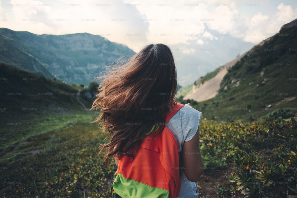 Young woman with waving long hair and backpack traveling along beautiful mountain meadows