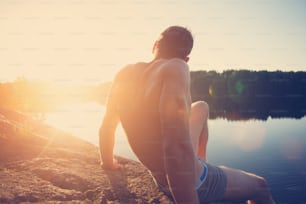 Joven musculoso sentado en el acantilado cerca del lago y mirando a lo lejos al atardecer (resplandor intencional del sol y destellos de lente)