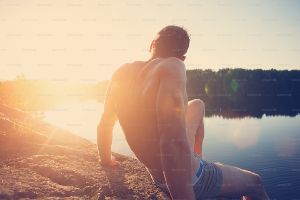 Jovem musculoso sentado no penhasco perto do lago e olhando para longe ao pôr do sol (brilho intencional do sol e reflexos de lente)