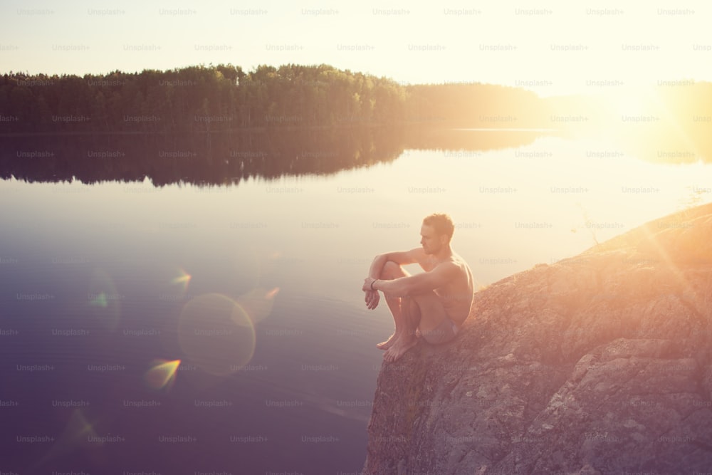 Jovem sentado na beira do penhasco perto do lago depois de nadar (brilho intencional do sol e reflexos das lentes)
