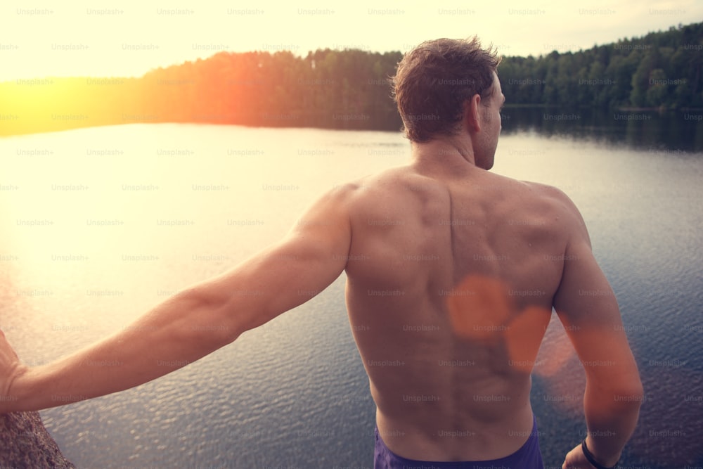 Homem jovem e musculoso de pé no penhasco perto da água ao ar livre e olhando para longe (brilho intencional do sol e reflexos de lentes)
