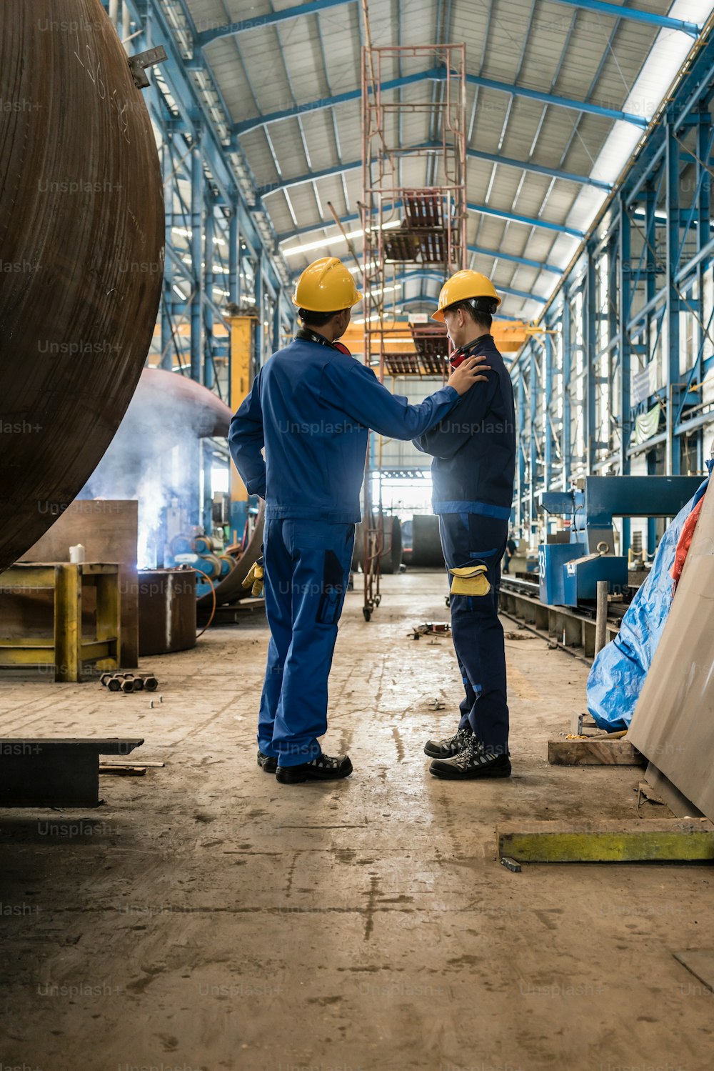 Two workers wearing yellow hard hat and blue uniform in the interior of an industrial hall