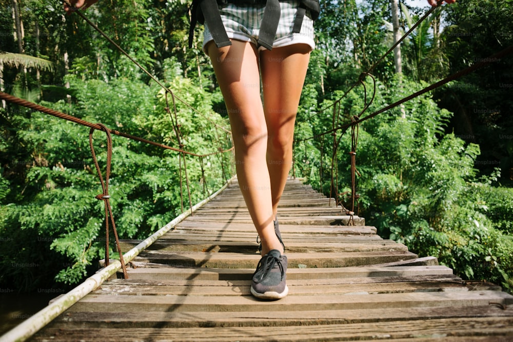 Pernas da mulher com mochila andando através da ponte suspensa na floresta tropical