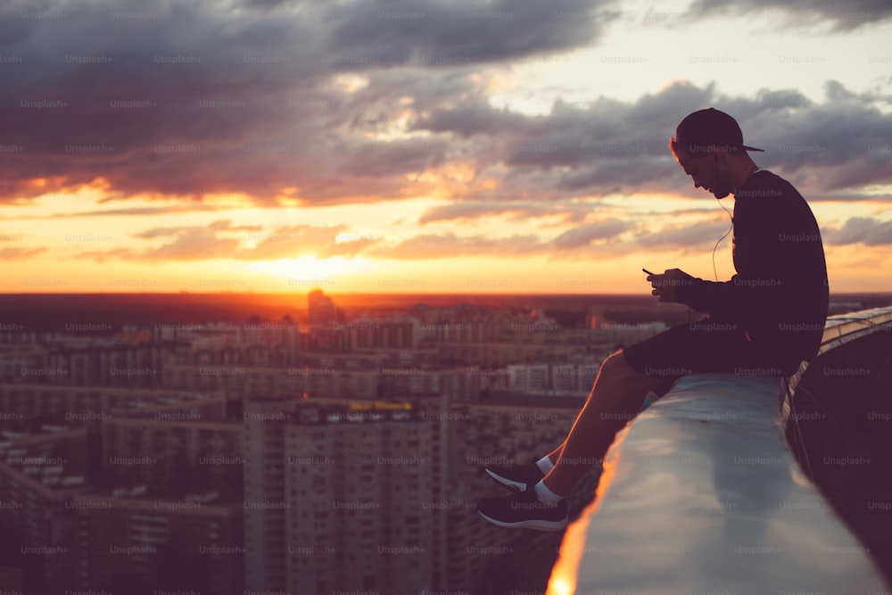 Jeune homme courageux assis au-dessus de la ville avec un smartphone au coucher du soleil