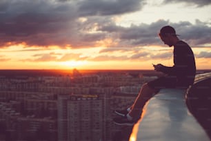 Jovem corajoso sentado acima da cidade com smartphone ao pôr do sol