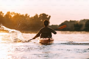 Rear view of young man splashing water while kayaking on river with sunset in the background