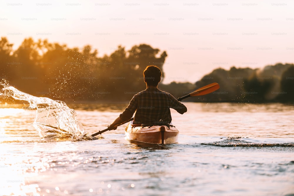 Rückansicht eines jungen Mannes, der Wasser spritzt, während er auf dem Fluss mit dem Kajak fährt, mit Sonnenuntergang im Hintergrund