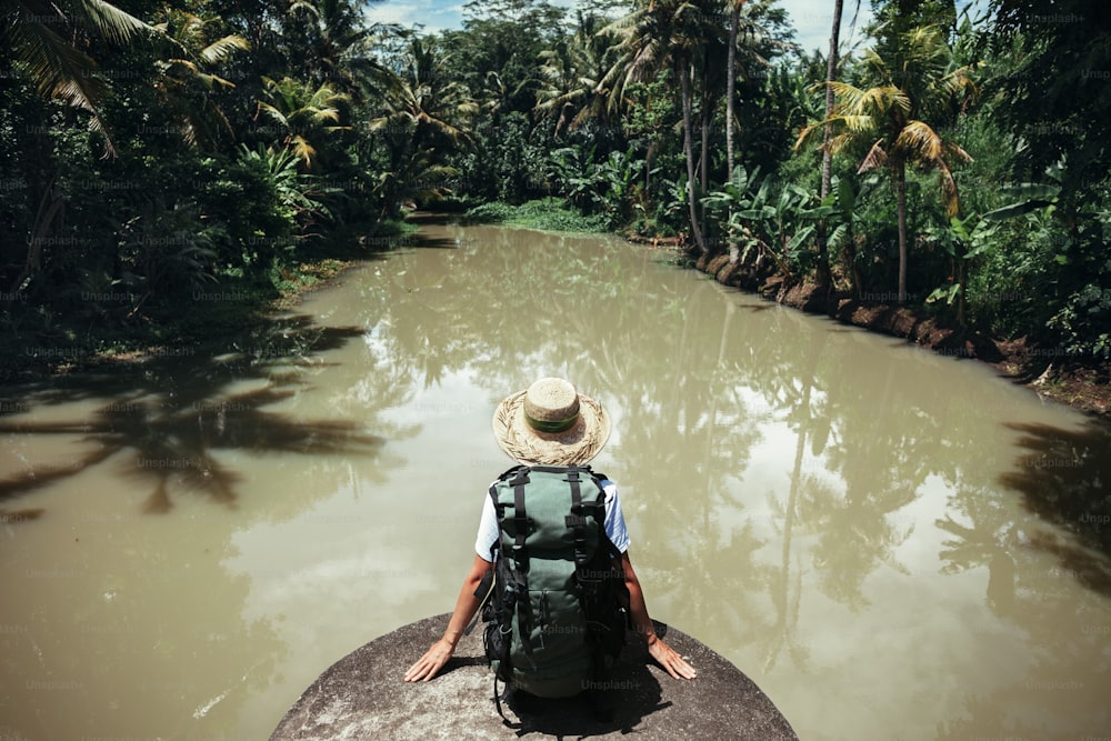 Mujer viajera joven sentada con mochila grande y mirando el río tropical