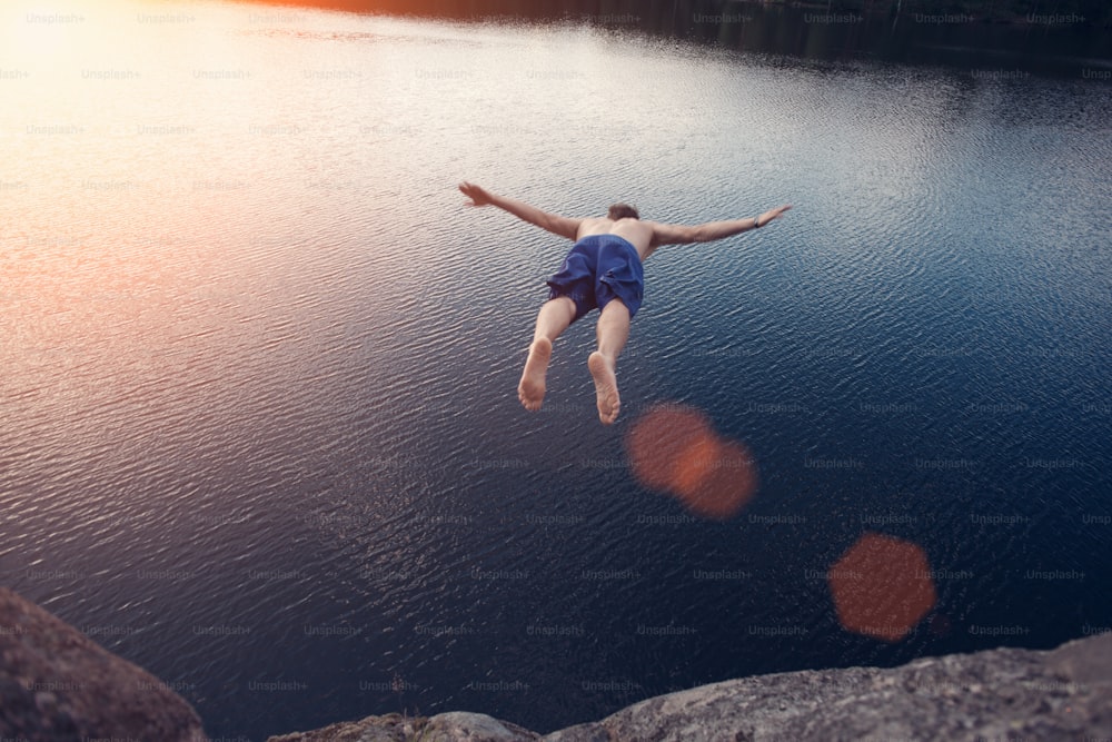 Jeune homme sautant dans l’eau depuis une falaise au coucher du soleil avec les mains écartées (éblouissement intentionnel du soleil et reflets de l’objectif, homme en flou de mouvement)