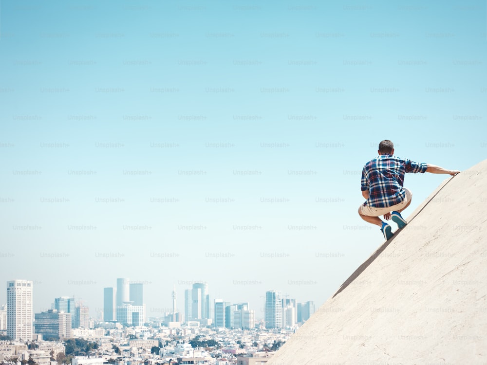 Hombre joven y arriesgado sentado en un pico y mirando la ciudad