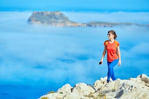 sporty female traveler with backpack standing on the cliff against sea and blue sky with white clouds at early morning, Crete, Greece. woman holding the phone smiling and looking at left side