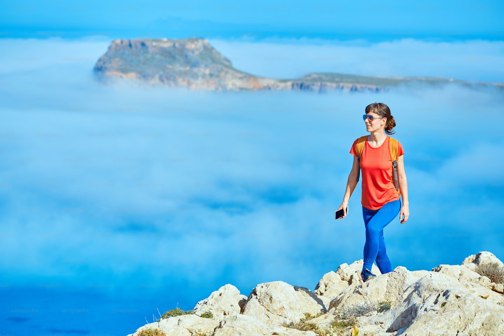 sporty female traveler with backpack standing on the cliff against sea and blue sky with white clouds at early morning, Crete, Greece. woman holding the phone smiling and looking at left side