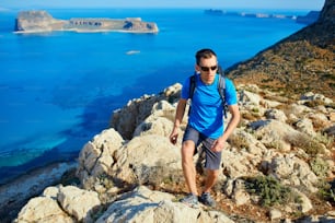 male traveler with backpack runs on the cliff against sea and blue sky at early morning. Balos beach on background, Crete, Greece