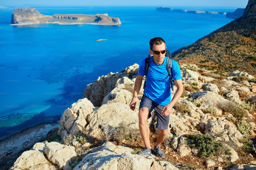 Un viajero masculino con mochila corre por el acantilado contra el mar y el cielo azul a primera hora de la mañana. Playa de Balos al fondo, Creta, Grecia