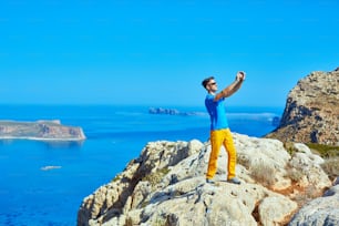이른 아침에 바다와 푸른 하늘을 배경으로 절벽에 서 있는 남자 여행자. 배경에 발로스 해변, 크레타, 그리스. 스마트폰으로 사진을 찍는 남자
