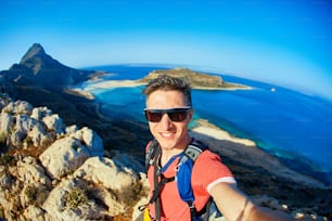 早朝の海と青空を背景に崖の上に立つリュックサックを背負った男の旅人。背景にバロスビーチ、クレタ島、ギリシャ。自撮りをする男