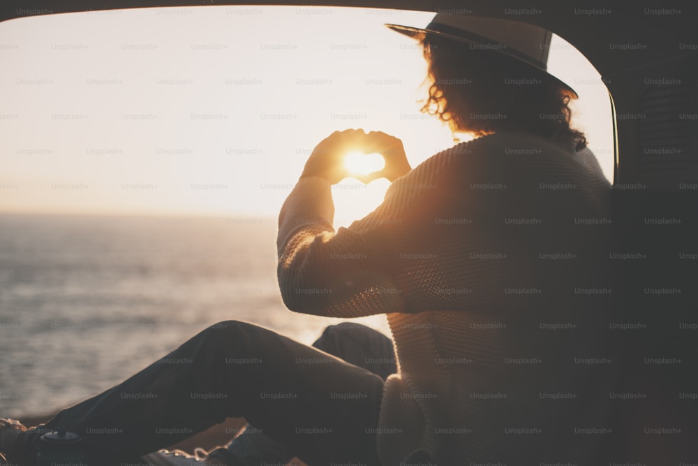 Young traveling female sitting in hat in the trunk of van and enjoing the adventure, woman hipster making heart symbol with hands