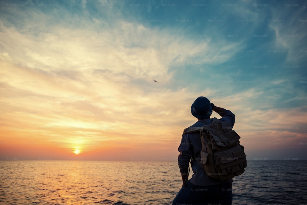 Turista con mochila cerca del mar mirando a lo lejos en el horizonte al atardecer