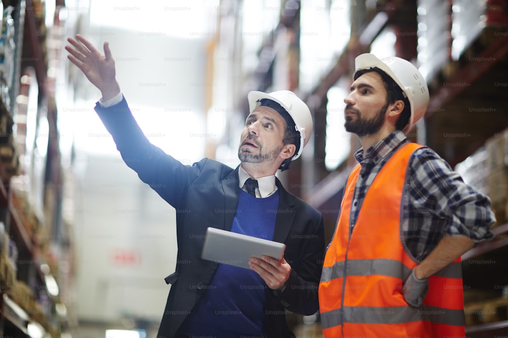 Businessman with touchpad pointing at upper shelf with goods while talking to worker