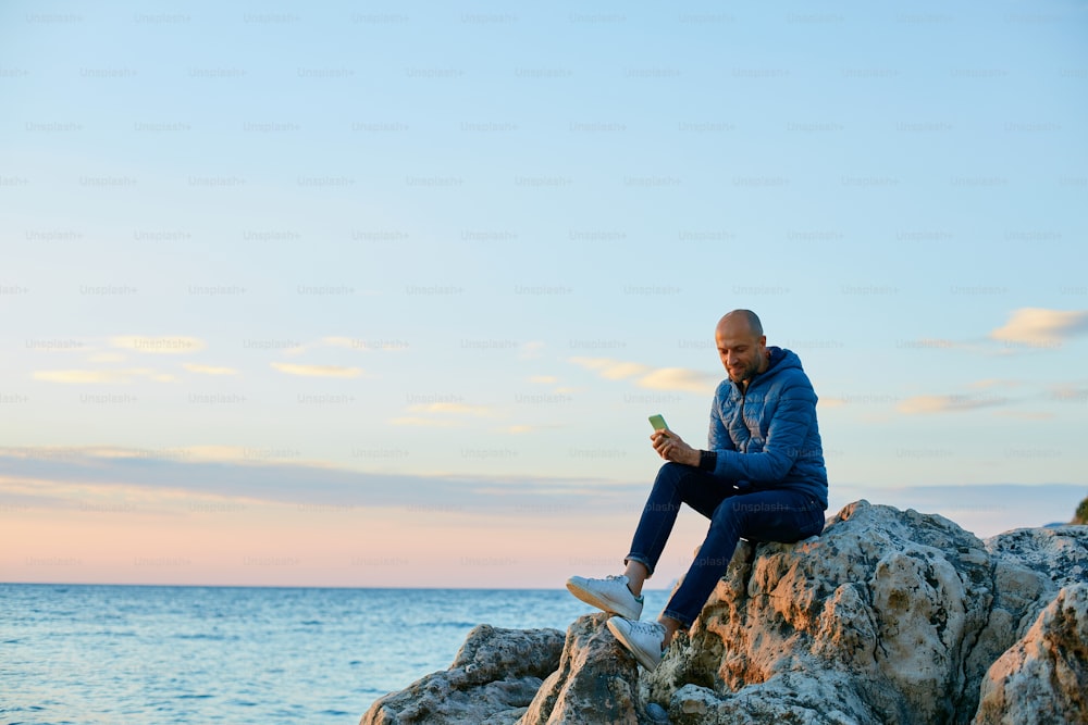 Hombre joven en la playa contra el mar y el sol de la salida del sol temprano en la mañana y mira el teléfono