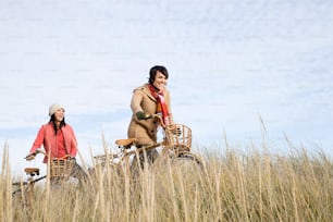 a couple of women riding bikes through a grass covered field