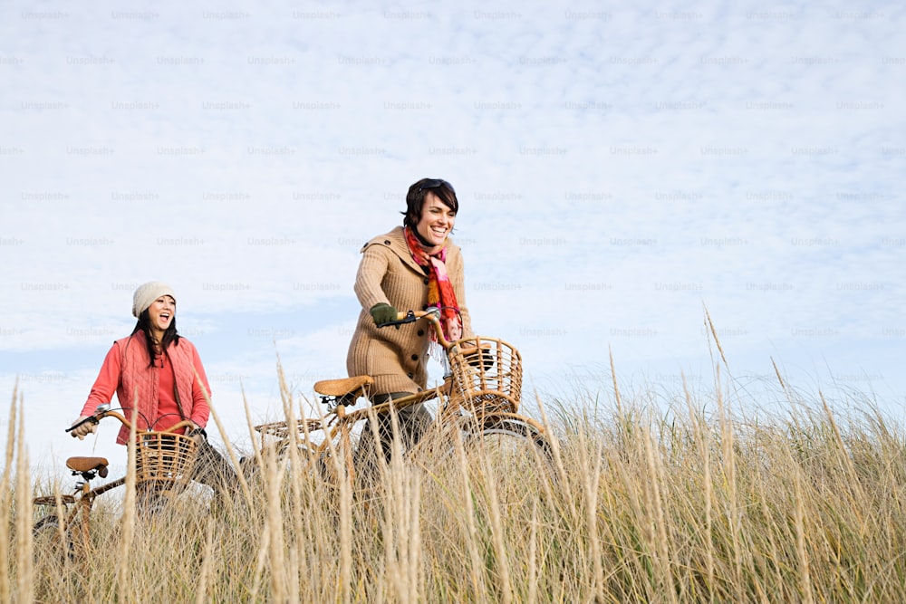 a couple of women riding bikes through a grass covered field