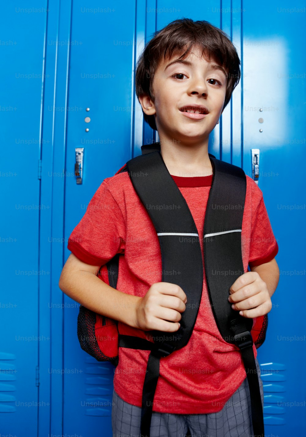 Retrato de un niño feliz de la escuela sosteniendo su mochila mientras está de pie frente al casillero