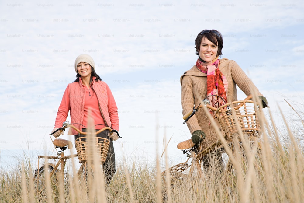 a couple of women riding bikes through a grass covered field