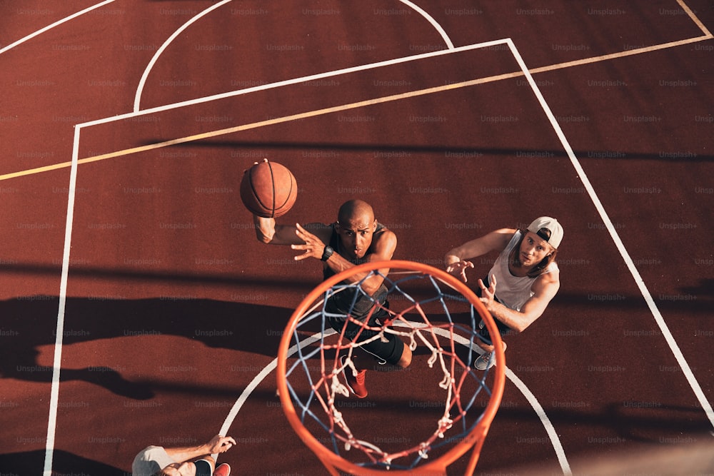 Vista superior de un joven con ropa deportiva anotando una volcada mientras juega baloncesto con amigos al aire libre