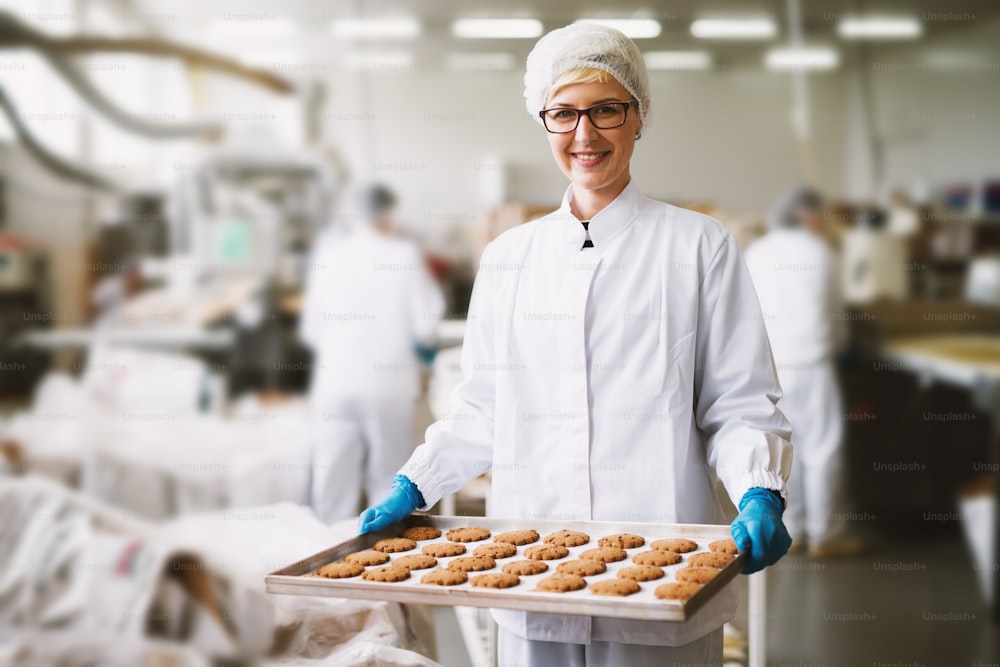 Young satisfied female worker in sterile clothes holding fresh made cookies in food factory.