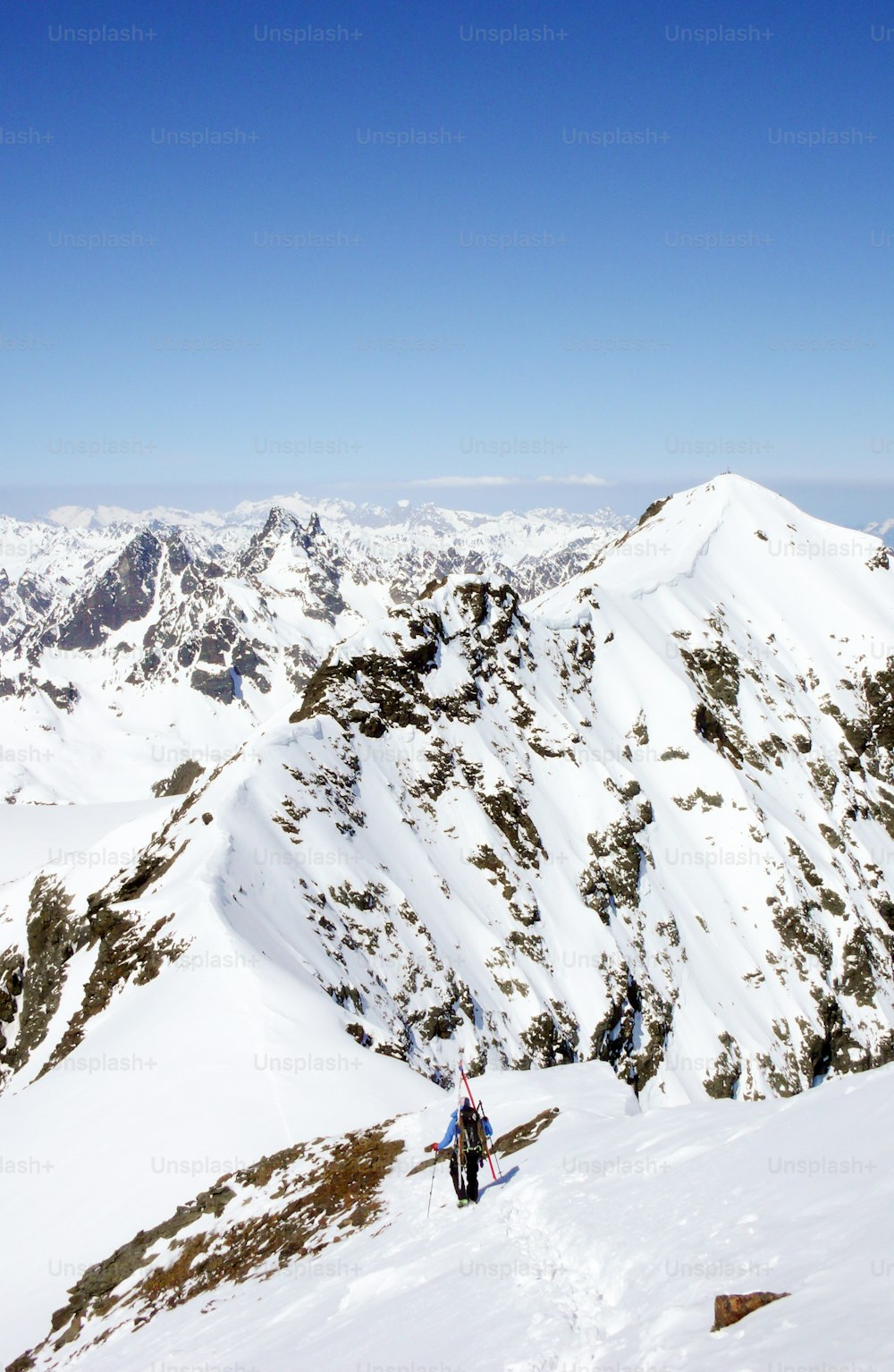 A male backcountry skier climbs and hikes a long exposed narrow snow ridge in the Alps of Switzerland on his way to a high mountain peak in the Silvretta mountain range.