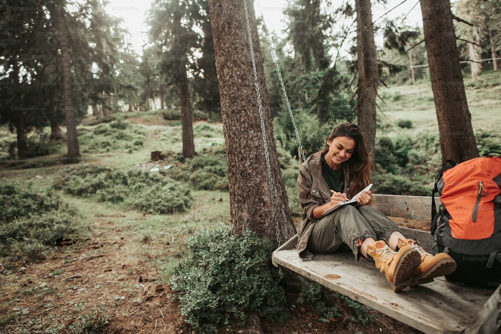 Esto es importante. Retrato de una hermosa joven escribiendo en un cuaderno y sonriendo. Árboles y plantas verdes en el fondo