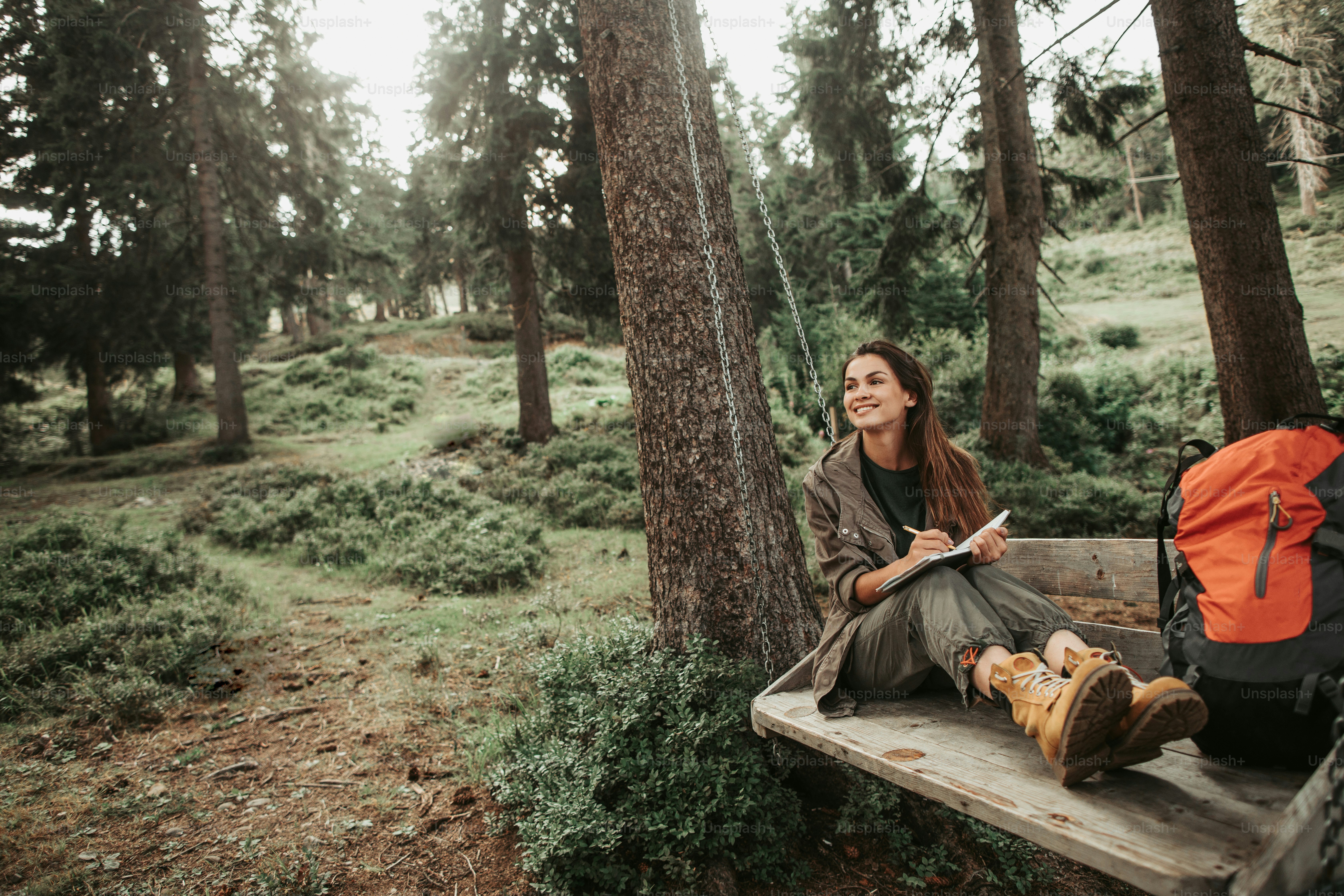 I like this place. Portrait of charming young lady holding notebook with pen while looking away with smile. Trees and green plants on background