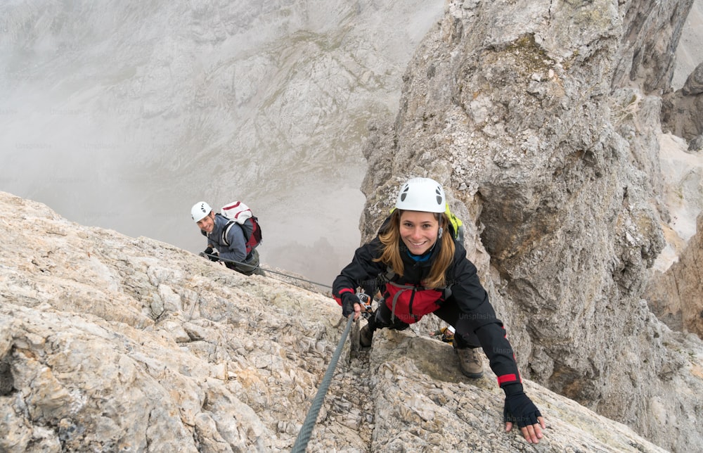 jovens alpinistas atraentes em uma rocha vertical e exposta escalam uma Via Ferrata enquanto sorriem e apontam para um pico de montanha distante