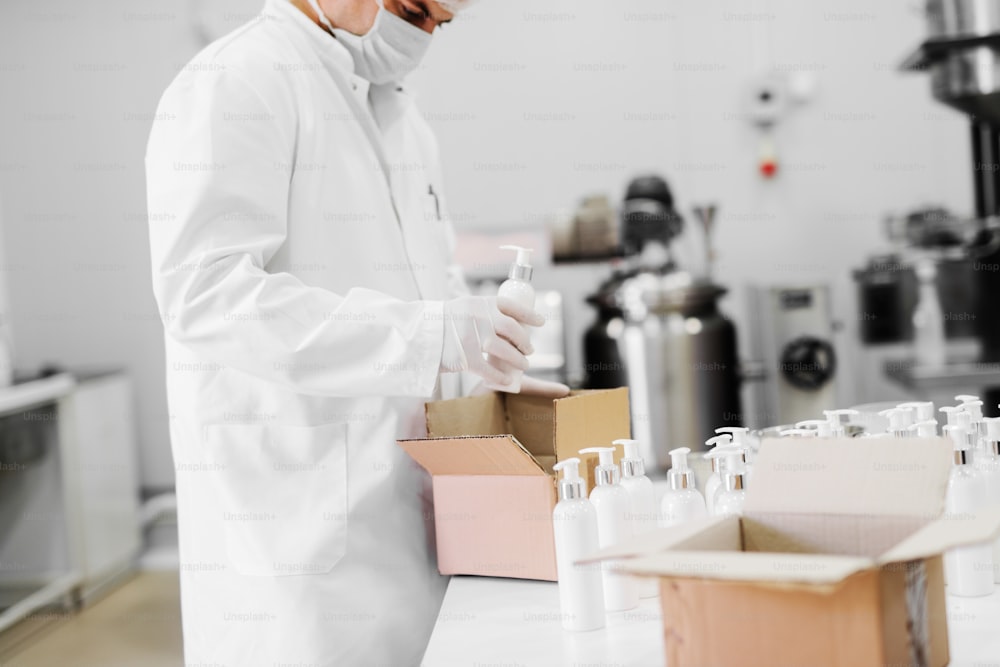 Imagen de un hombre con ropa estéril empacando botellas con loción en caja de cartón. De pie en el laboratorio brillante y empaquetando productos.