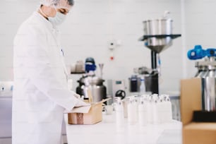 Bild eines Mannes in steriler Kleidung, der Flaschen mit Lotion im Karton verpackt. Stehen in hellen Labor- und Verpackungsprodukten.