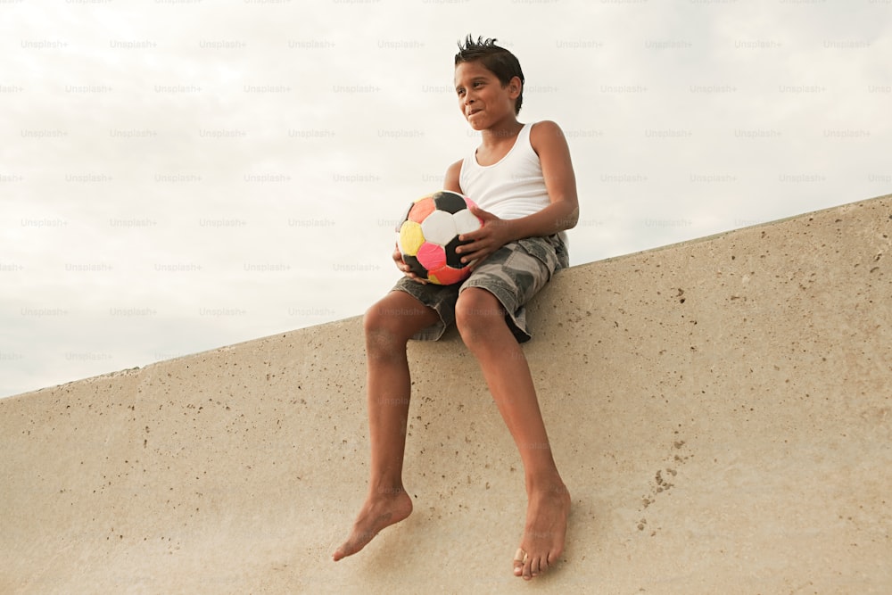 a boy sitting on a wall holding a soccer ball
