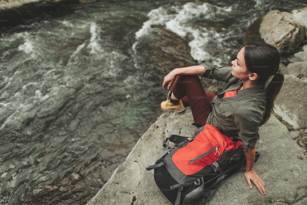 Jeune femme ravie assise sur la rive de la rivière de pierre tout en ayant un week-end actif