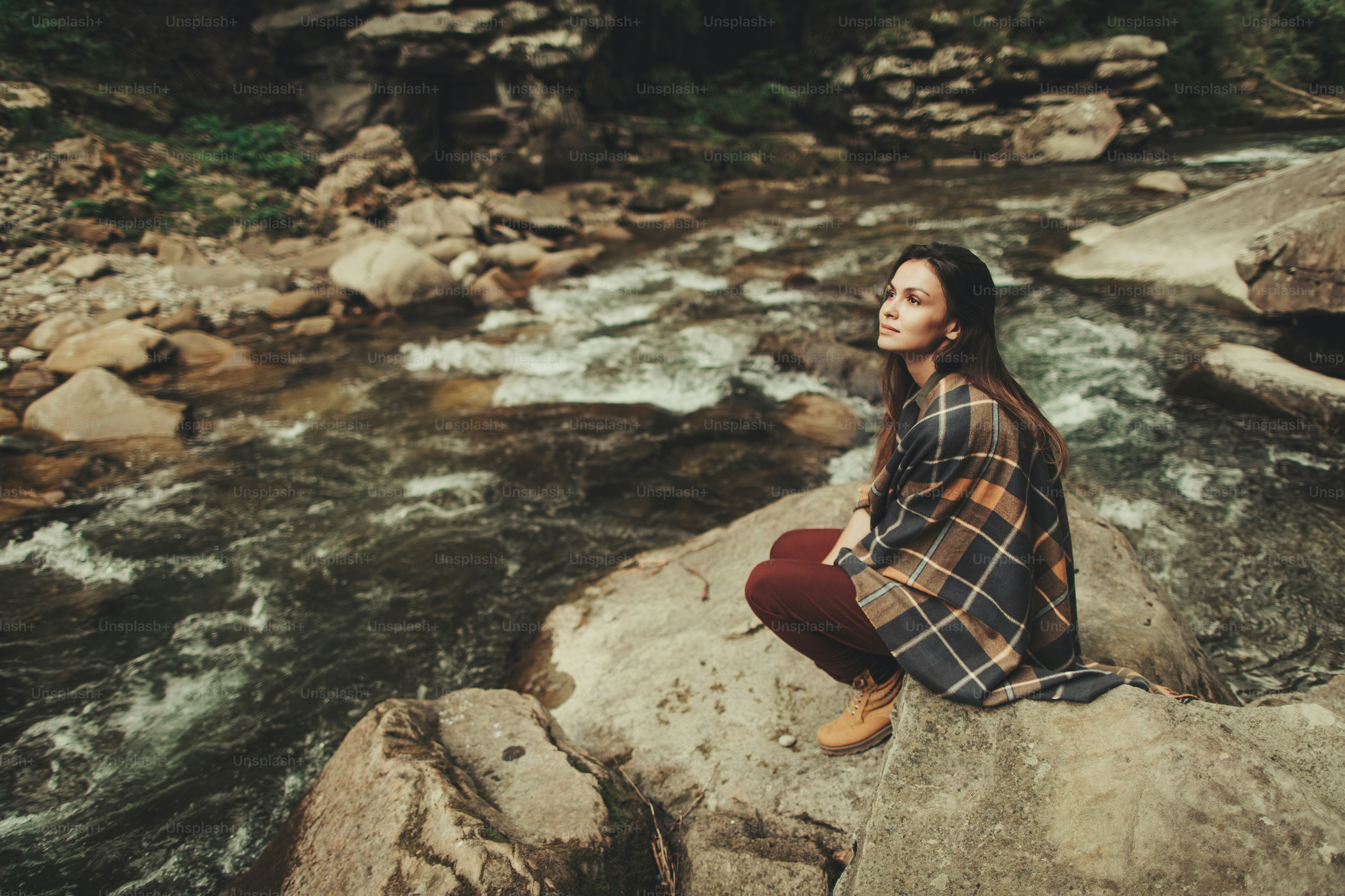 Pleased young woman being involved in thoughts while sitting on the bank of mountain river