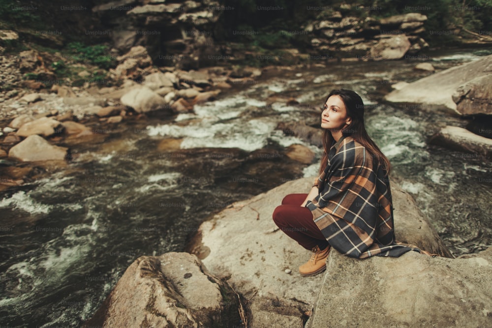 Mujer joven complacida que está involucrada en pensamientos mientras está sentada en la orilla del río de la montaña