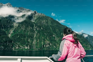 젊은 여성 관광객이 뉴질랜드 남섬 피오르드랜드 국립공원의 밀포드 사운드에 있는 선박 갑판에서 피요르드 풍경을 바라보고 있다. 밀포드 사운드의 보트 크루즈는 대부분의 관광객에게 주요 활동입니다.