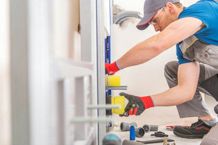 Plumber in a cap and safety glasses fitting pipe inside an open wall