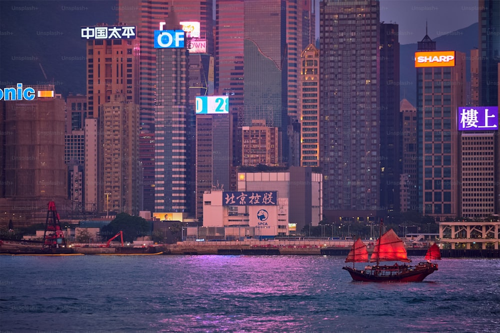 香港、中国 - 2018年5月1日:夕方にビクトリアハーバー上の赤い帆と香港のスカイラインの街並みのダウンタウンの高層ビルと観光ジャンクボートフェリー。香港、中国