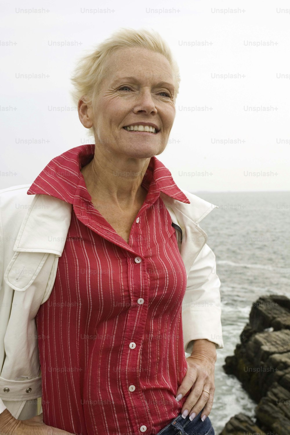 a woman in a red shirt and white jacket by the ocean